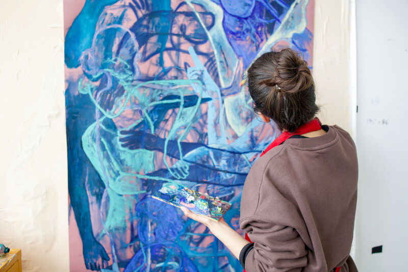 A person with long brown hair in a bun, painting in blues, pinks and purples on a large canvas.